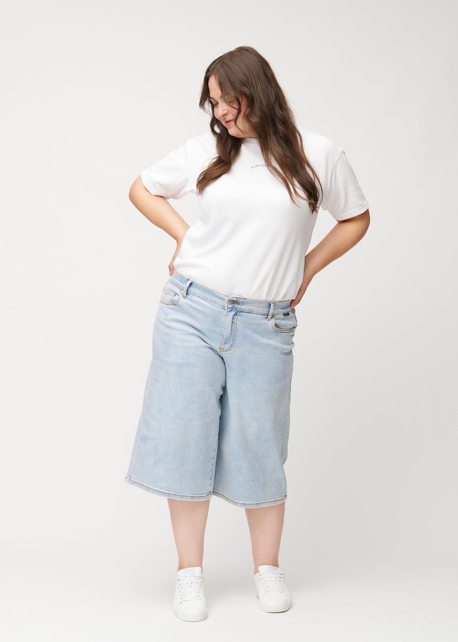 Fuldt billede af plus-size model i løse, lyseblå denim capris i farven Waves, der går til lige under knæet.