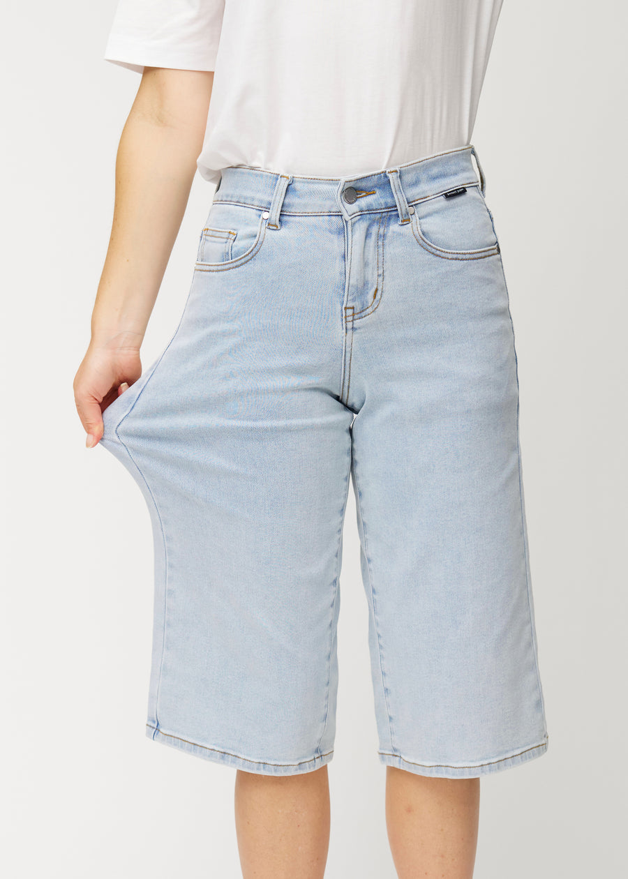 Model strækker de løse, lyseblå denim capris i farven Waves ved hoften for at vise materialets stretch.