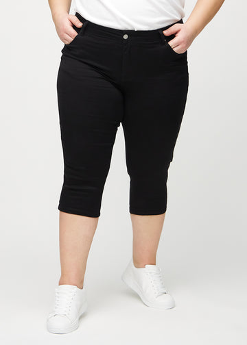 Black skinny capris, model name Ravens, which fit closely to the leg and end just below the knee on a plus-size model, seen from the front.