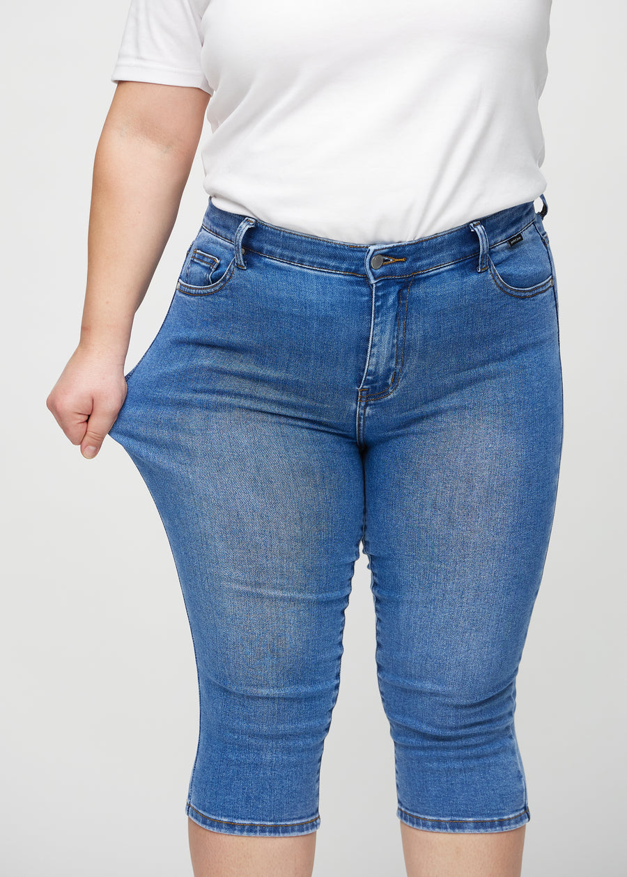 Skinny capris for women in blue denim, color Rivers. A plus-size model stretches the fabric to demonstrate its flexibility.