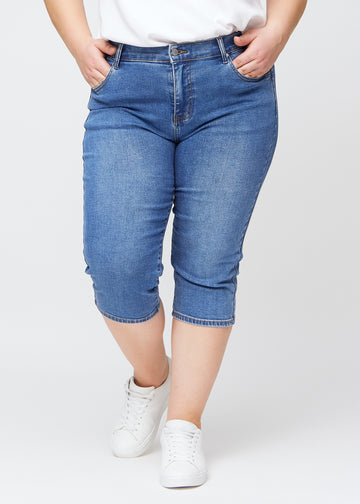 Slim capris for women in blue denim, color Rivers. Front view on a plus-size model, showing the fitted cut ending just below the knee.
