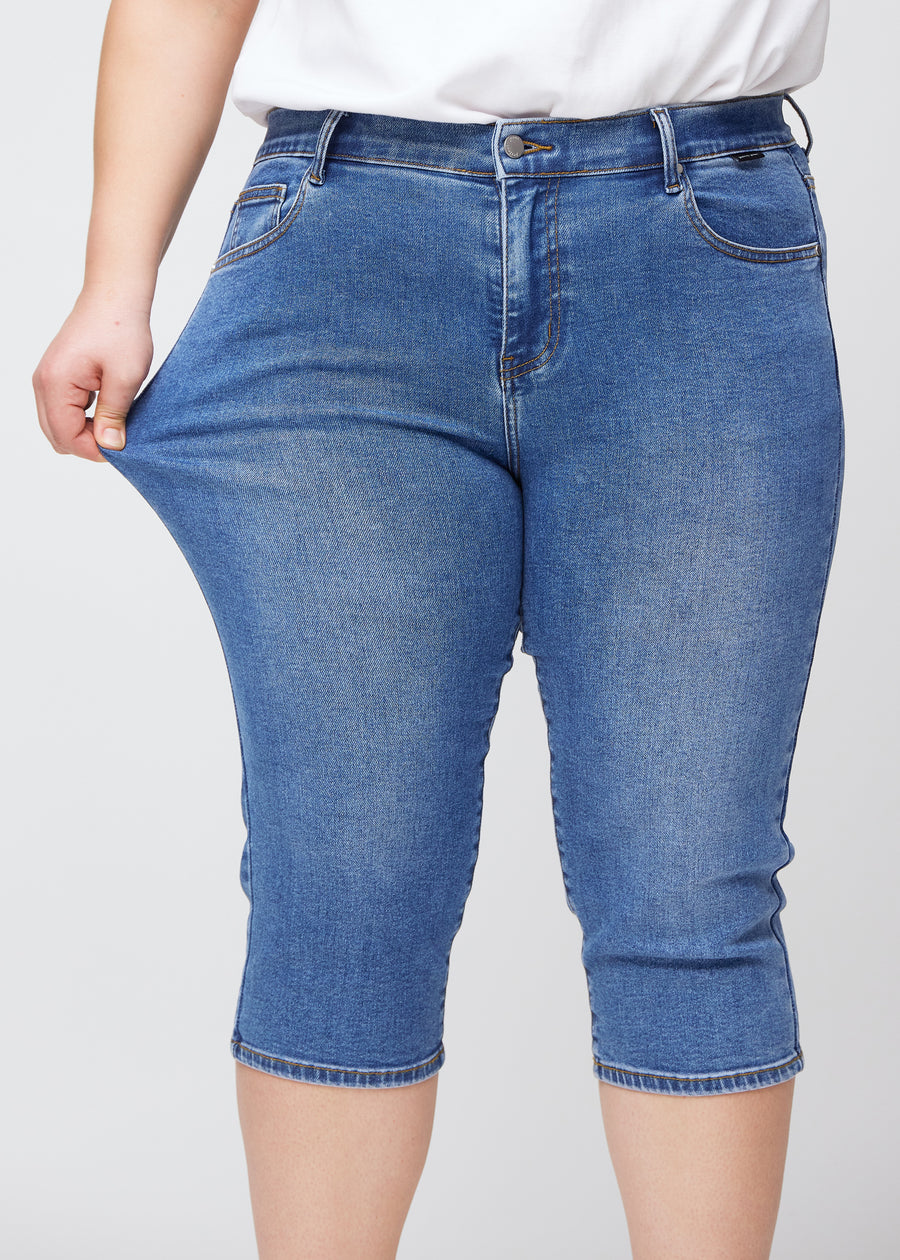 Slim capris for women in blue denim, color Rivers. A plus-size model pulls the fabric at the thigh to demonstrate stretch and flexibility.