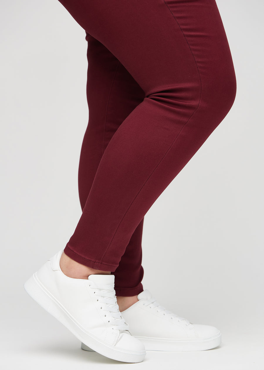 Skinny jeans for women in dark red, color Santas. Close-up of the snug-fitting leg openings at the ankles on a plus-size model.
