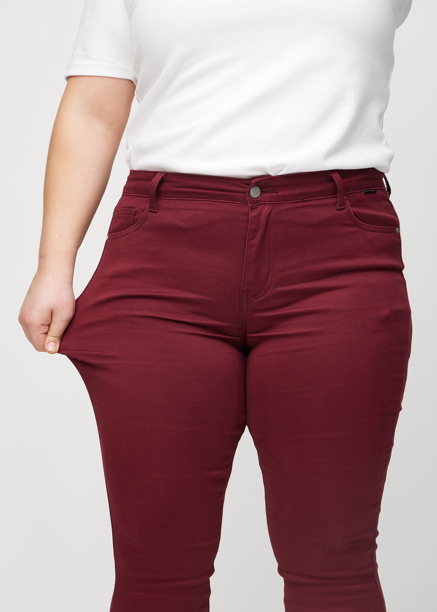 Skinny jeans for women in dark red, color Santas. A plus-size model stretches the jeans at the thigh to show the fabric&