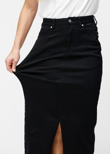 Model strækker den lange, sorte jeansnederdel i farven Ravens ved hoften for at vise materialets stretch.